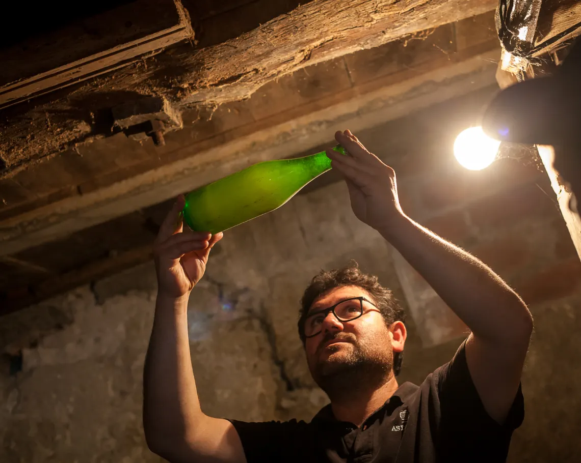 An Astarbe cider maker holds a green cider bottle up to the light in the stone and timber cellar