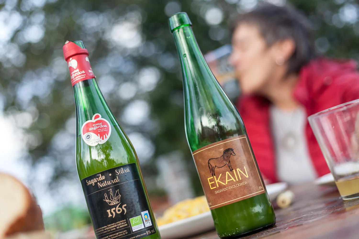 Two bottles of Ekain organic cider on an outdoor table, with someone drinking in the background