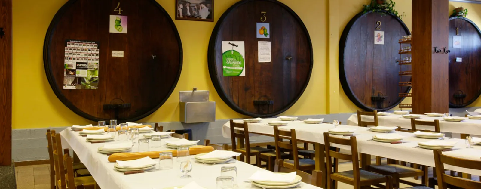 Comedor de la sidrería Aginaga, con mesas largas de mantel blanco ante grandes kupelas numeradas