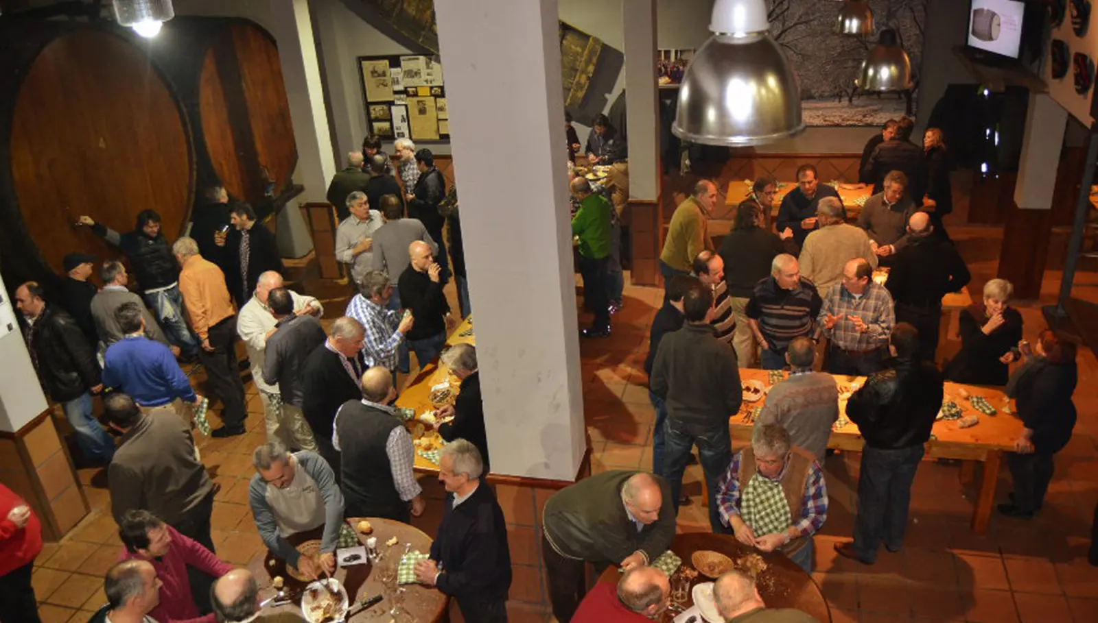 Comedor lleno de gente de pie junto a las mesas y las grandes kupelas de la sidrería Altzueta, visto desde arriba