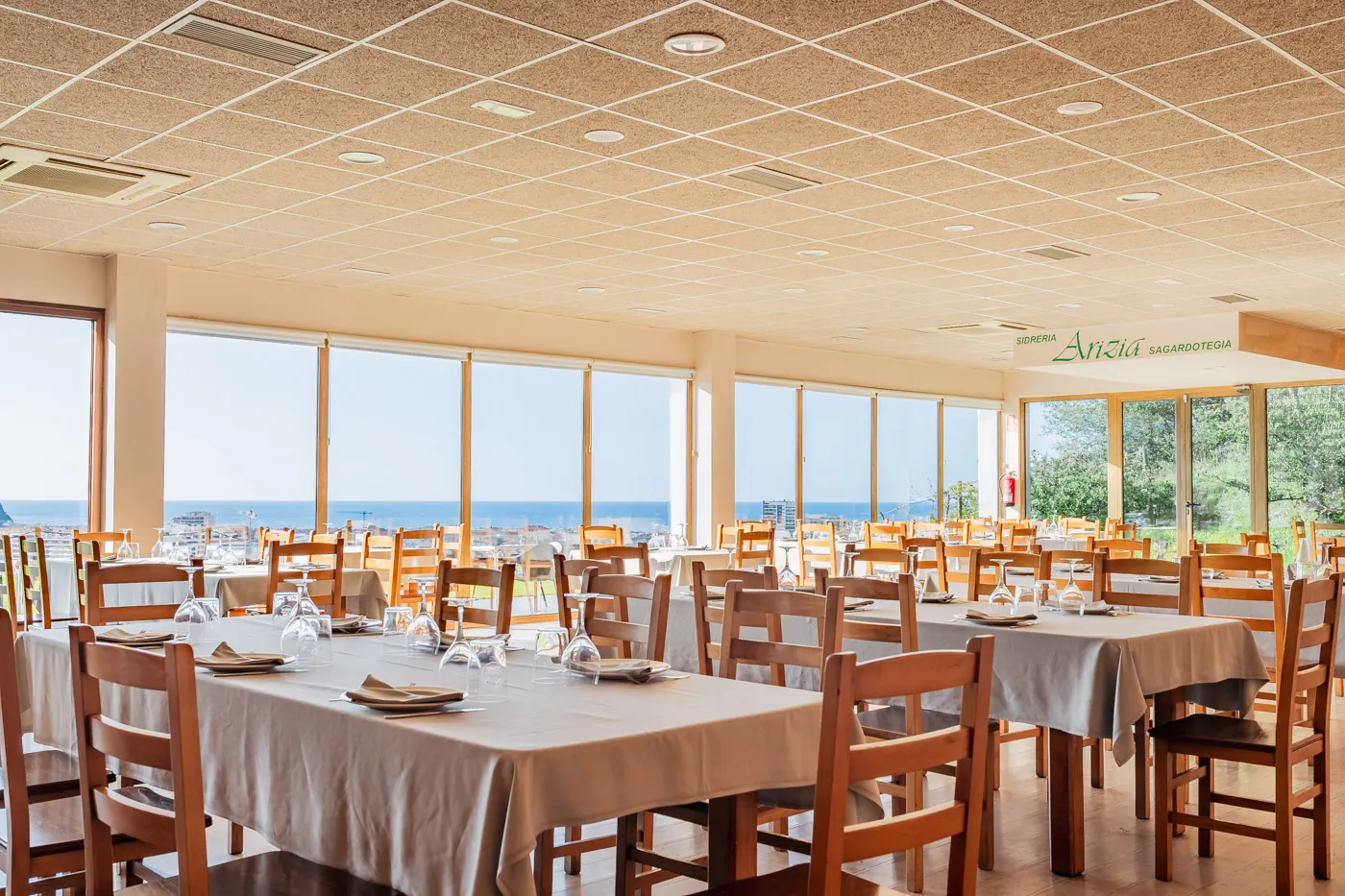 Comedor de la sidrería Arizia con las mesas preparadas y grandes ventanales con vistas al mar