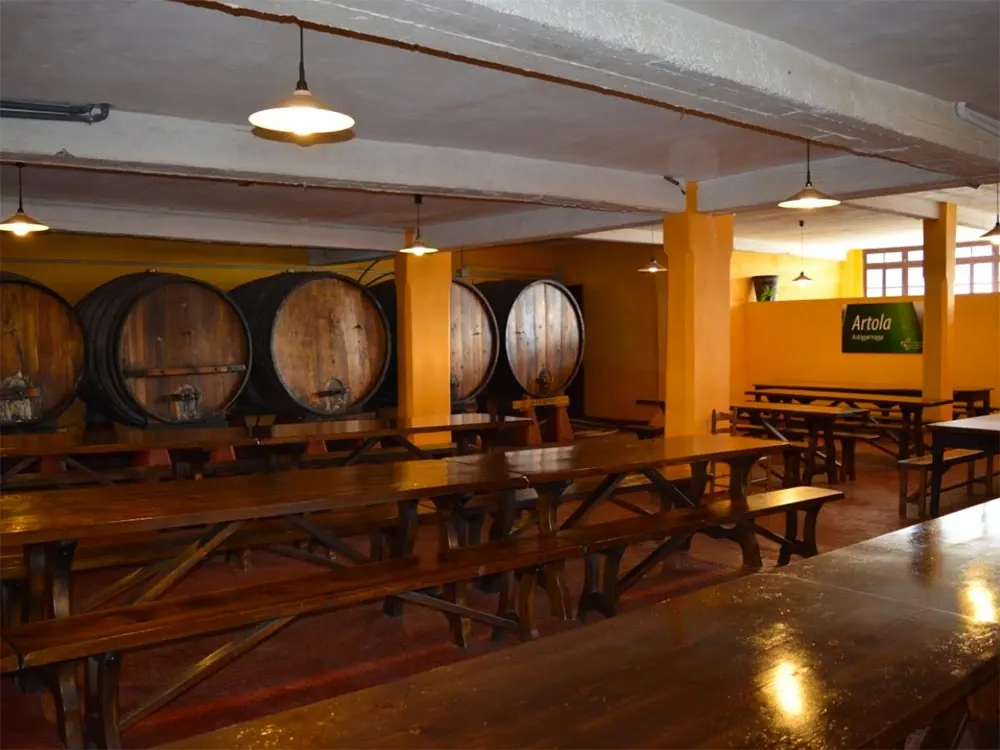 Comedor de la sidrería Artola, con mesas y bancos de madera, columnas amarillas y una fila de kupelas al fondo