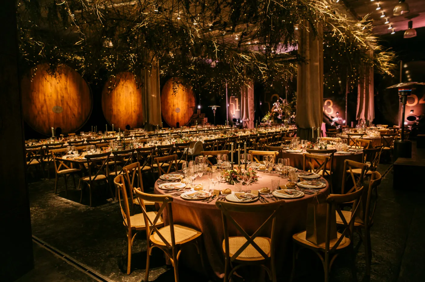 Mesas preparadas para un evento en un llagar de Casa Trabanco, con grandes toneles iluminados al fondo