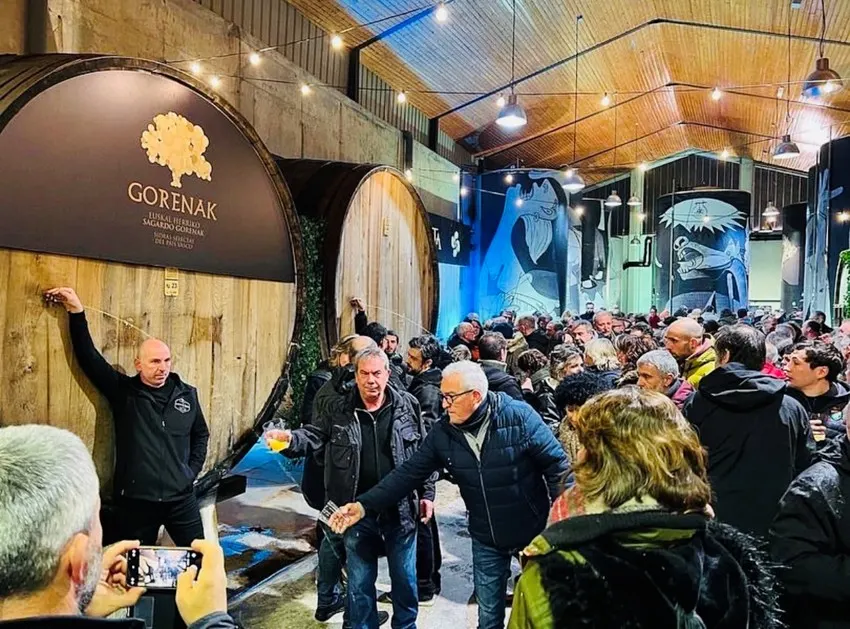 Txotx en la bodega de Gurutzeta, con la kupela de sidra Gorenak, gente llenando el vaso y murales en los depósitos