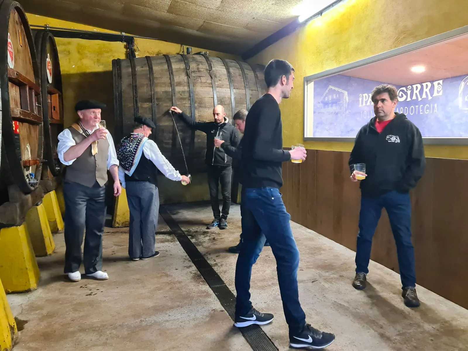 Txotx en la bodega de Iparragirre, con un hombre sacando sidra de una gran kupela y otros con el vaso en la mano