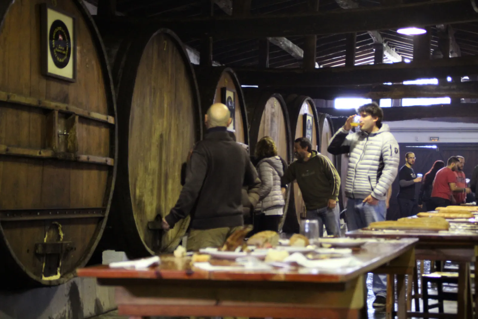 Txotx entre las grandes kupelas de la sidrería Iruin Astiazarán, con una mesa larga con pan y comida delante