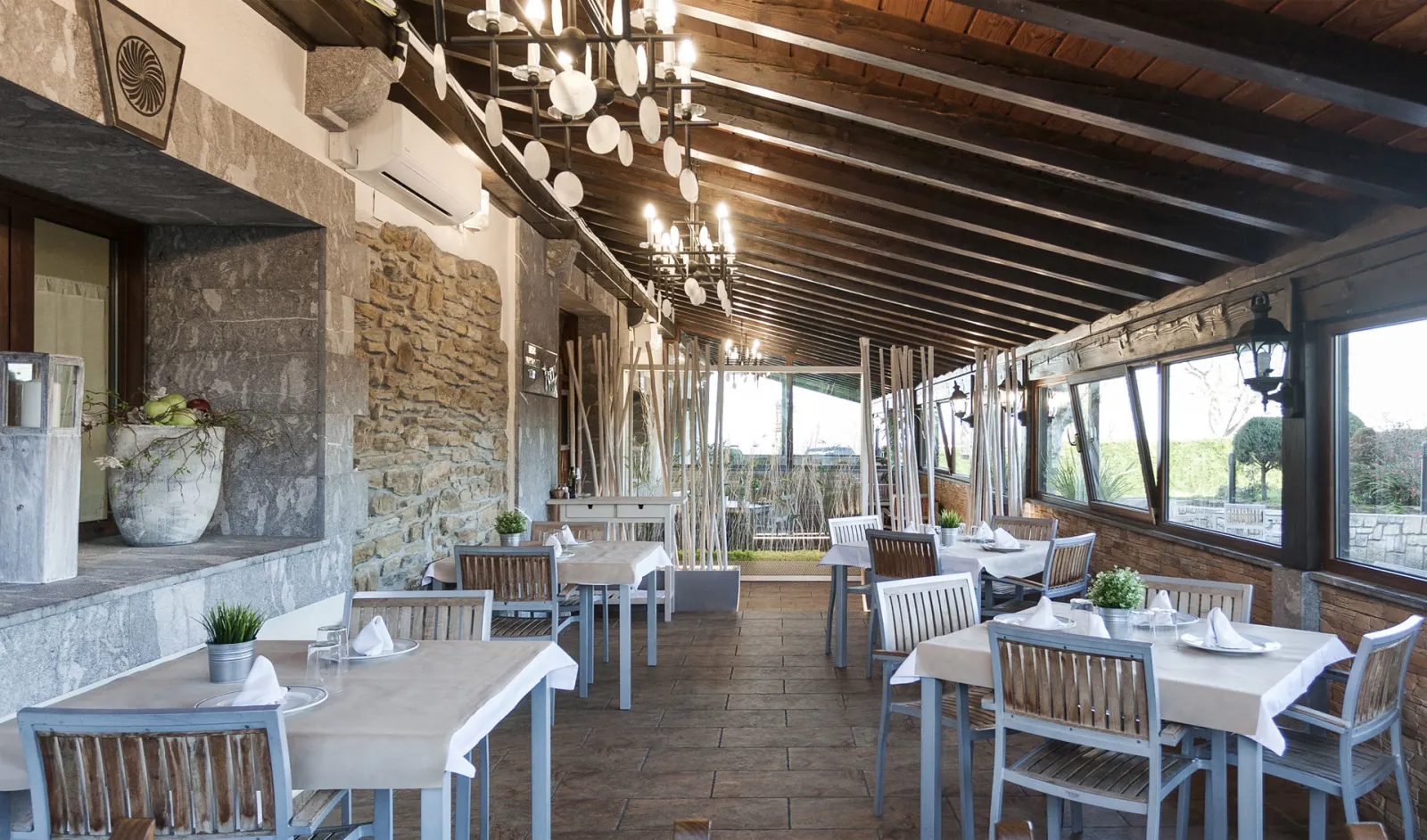 Comedor acristalado de la sidrería Izeta, con vigas de madera, pared de piedra y mesas con mantel blanco