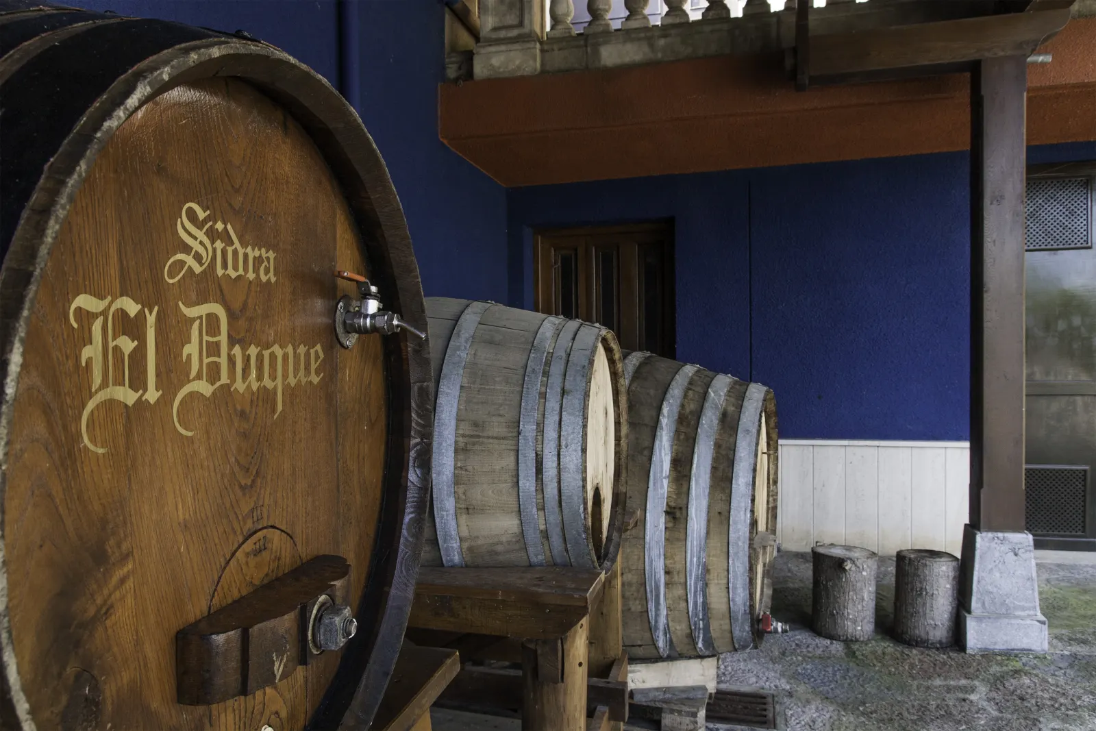 Barrica con el rótulo «Sidra El Duque» y otros toneles junto a la fachada azul del Llagar El Duque en Gijón