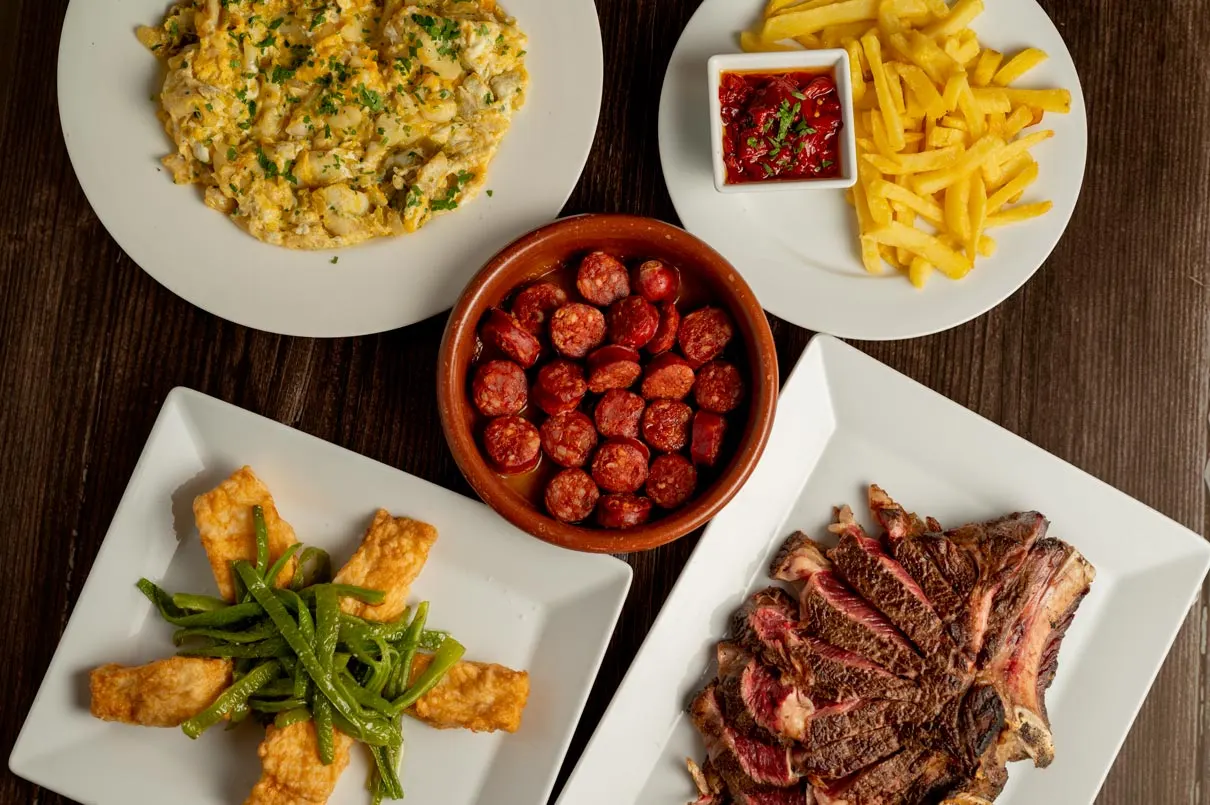 Dishes from the cider house menu at Loiu, seen from above, with chorizo in cider, cod scramble, battered cod with green peppers, T-bone steak and chips