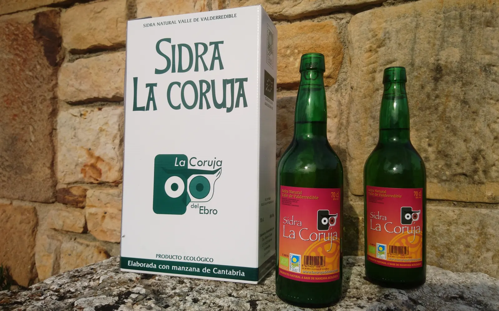 Caja y dos botellas de sidra natural ecológica La Coruja sobre un muro de piedra
