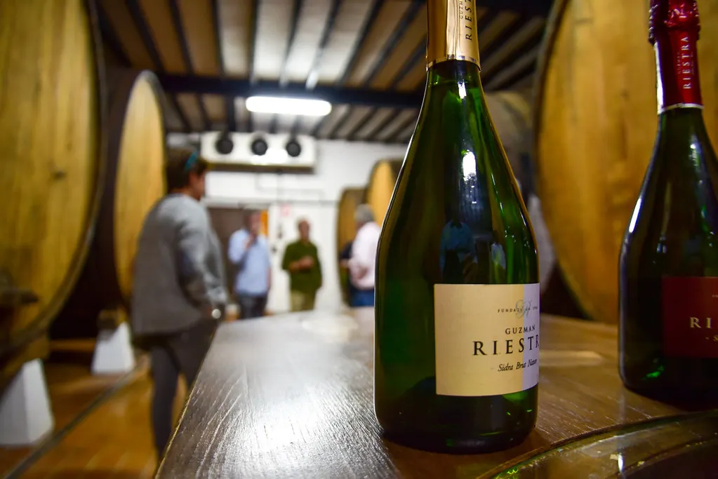 Bouteilles de cidre mousseux Riestra sur un tonneau, avec des personnes au fond du chai