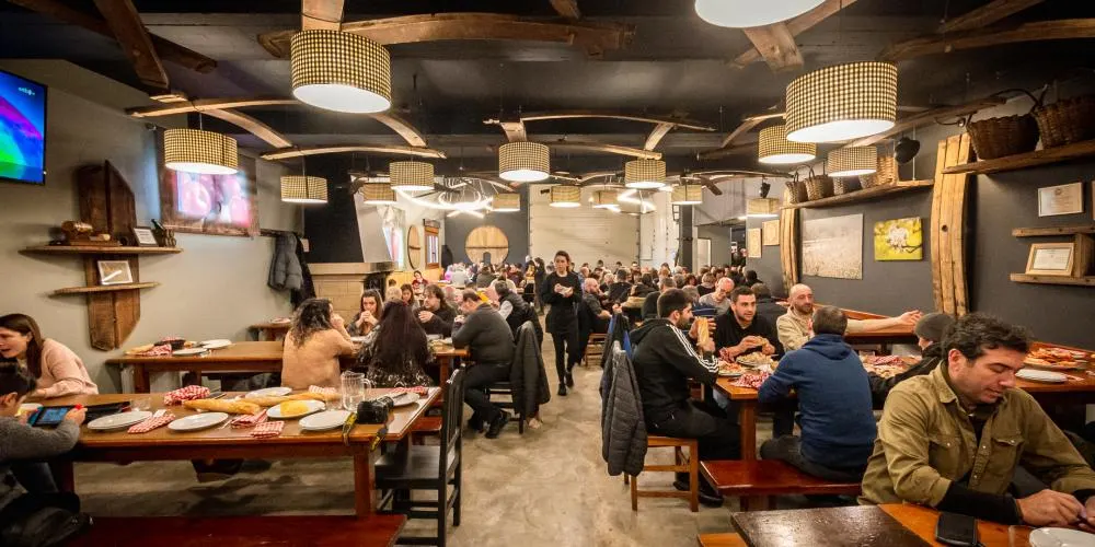 Comedor de la sidrería Urdaira lleno de comensales en mesas de madera, bajo lámparas de tela a cuadros