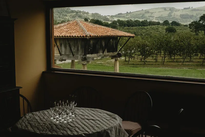Mesa con copas junto a un ventanal con vistas a un hórreo y a la pomarada